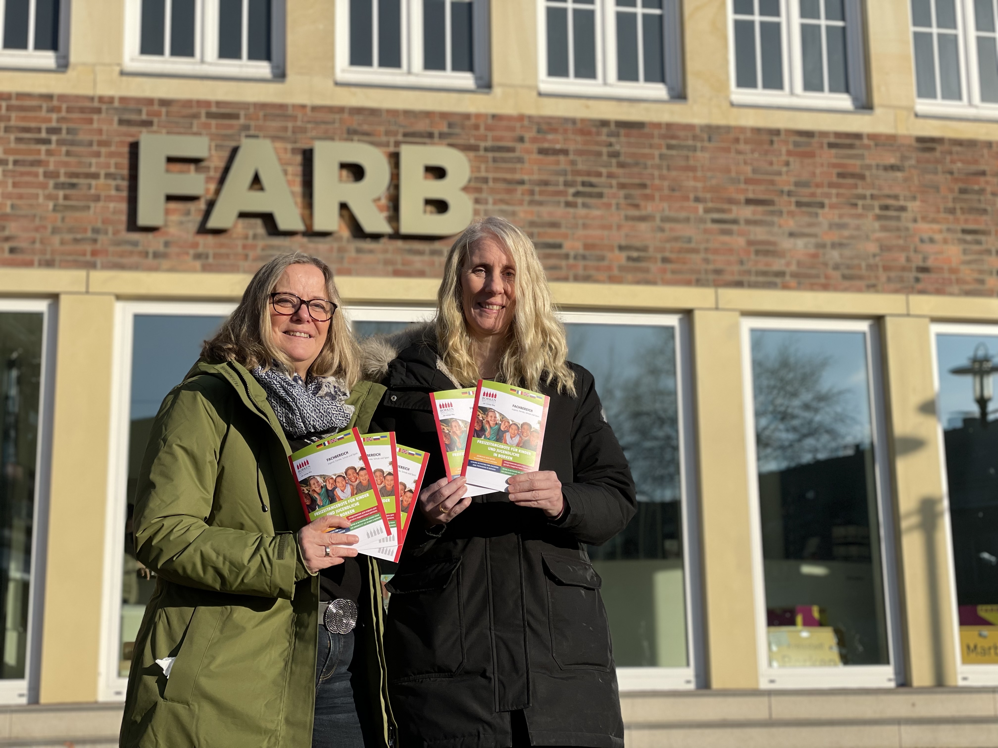 Die Jugendförderung der Stadt Borken stellt eine Übersicht über verschiedene Freizeitorte in verschiedenen Sprachen zur Verfügung. Sabine Sauret (re.) und Birgit Lütjann-Nienhaus von der Jugendförderung präsentieren den Freizeitflyer vor dem FARB. Dort, am Rathaus, in der Volkshochschule und in den Jugendhäusern im Stadtgebiet liegt der Flyer kostenlos aus.